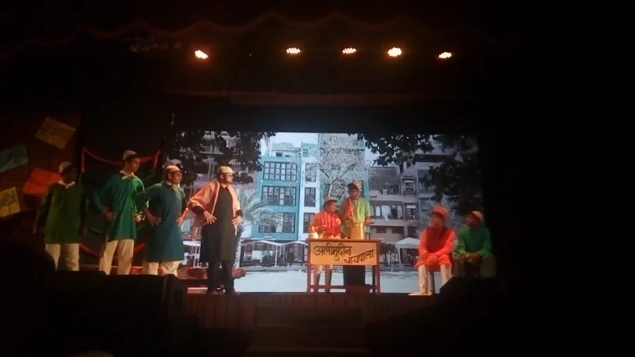 Actors in green costumes performing a play on a lit stage against a painted city backdrop