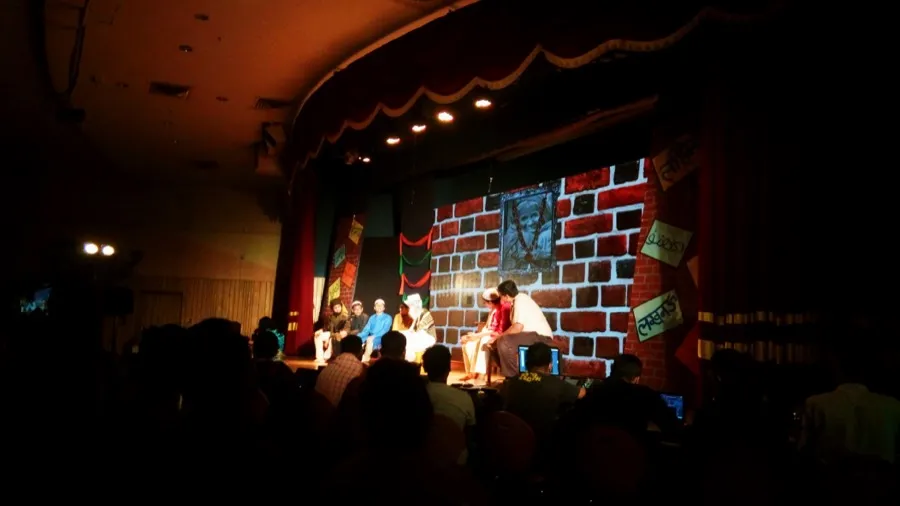 Audience watching a play staged against a painted brick wall backdrop